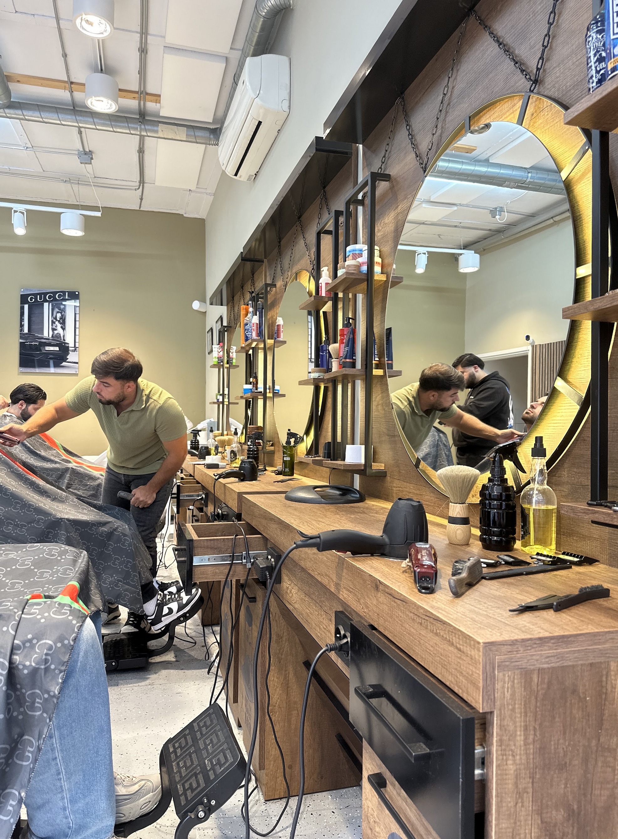 Interieur van Icon Barber: twee kappers aan het werk aan houten werkplekken met verlichte ronde spiegels
